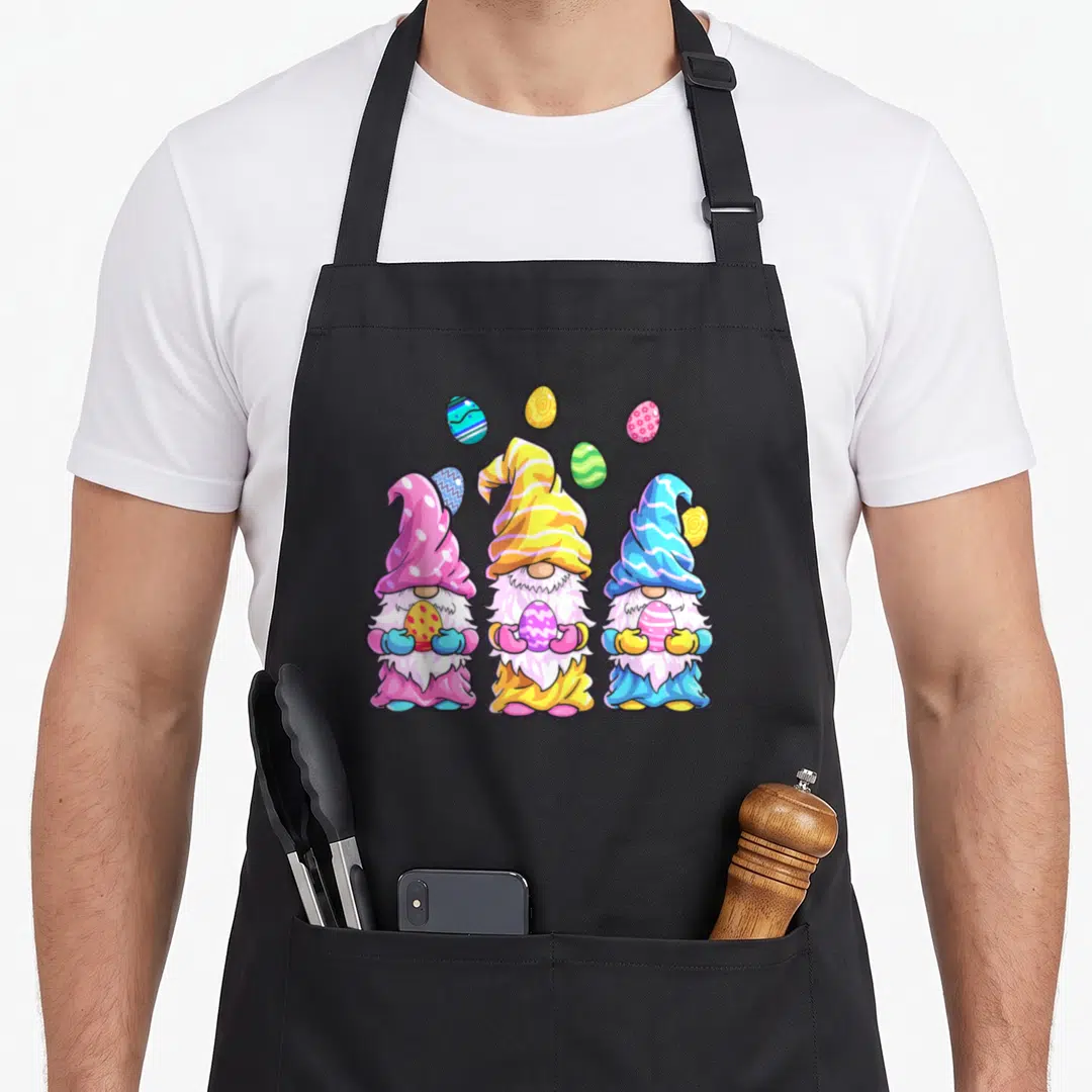 Outfit easter apron - Three cartoon gnomes holding eggs in pink, yellow, blue, and green with floating eggs above — shown on a black cotton canvas apron against a white background