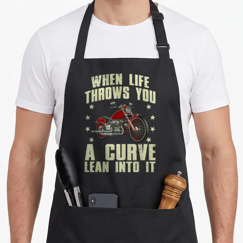 Funny racer motorcycle apron - Retro red motorcycle illustration flanked by stars with beige text when life throws you — shown on a black cotton canvas apron against a white background