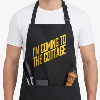 Cozy cottage apron - Yellow stacked text reading I'M COMING TO THE COTTAGE on black fabric — shown on a black cotton canvas apron against a white background