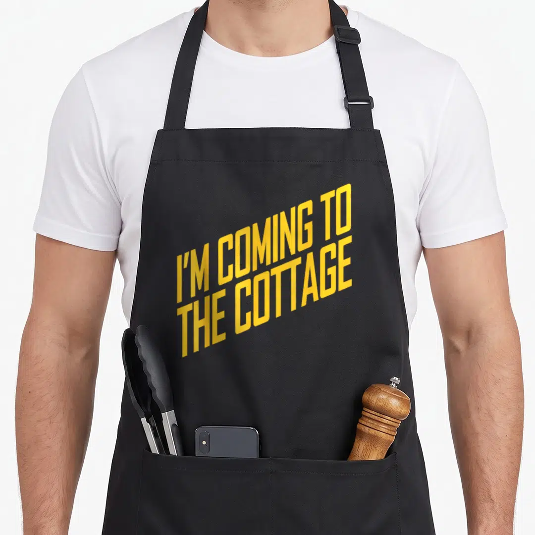 Cozy cottage apron - Yellow stacked text reading I'M COMING TO THE COTTAGE on black fabric — shown on a black cotton canvas apron against a white background