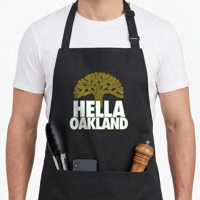 Hella tree apron - Yellow stylized tree above white HELLA OAKLAND text with green outlines — shown on a black cotton canvas apron against a white background