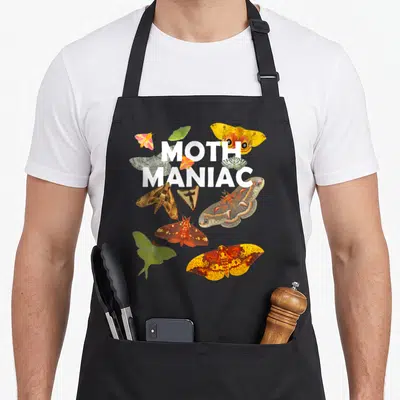 Maniac moth apron - White MOTH MANIAC text surrounded by colorful photo moths in yellow green brown red — shown on a black cotton canvas apron against a white background