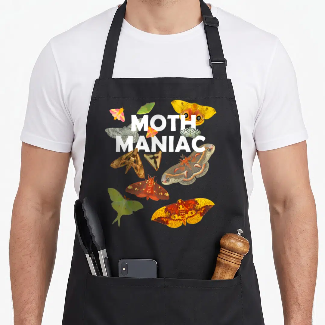 Maniac moth apron - White MOTH MANIAC text surrounded by colorful photo moths in yellow green brown red — shown on a black cotton canvas apron against a white background
