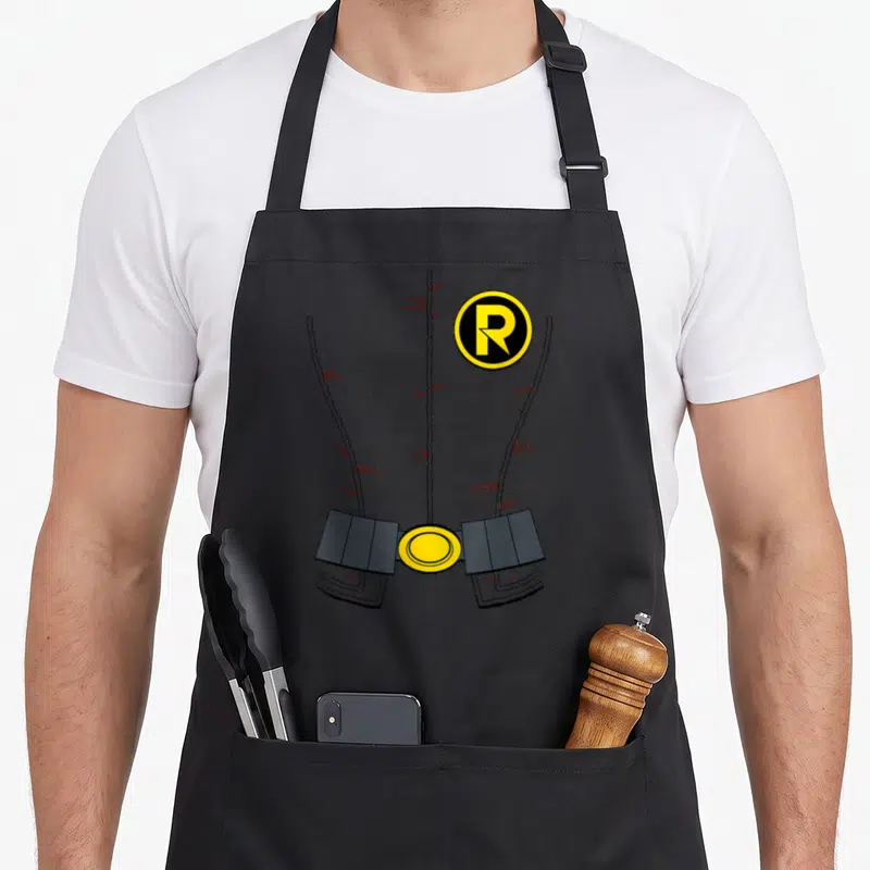 Robin uniform apron - Grey costume torso with yellow R logo, red scratch marks, and utility belt — shown on a black cotton canvas apron against a white background