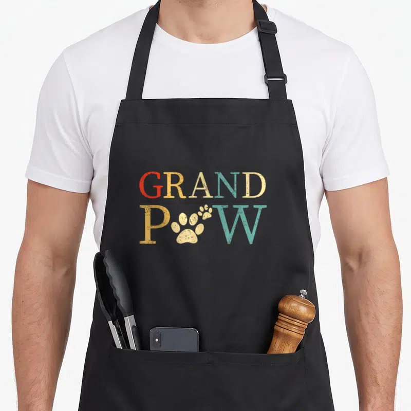 Papaw dog apron - Retro GRAND POW text with beige paw print replacing O in red yellow and teal letters — shown on a black cotton canvas apron against a white background