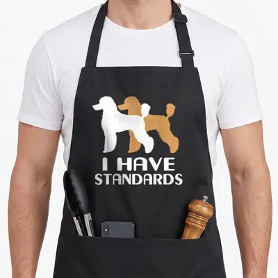 Toy poodle standard size of apron - Two overlapping poodle silhouettes in white and brown with stacked white text — shown on a black cotton canvas apron against a white background