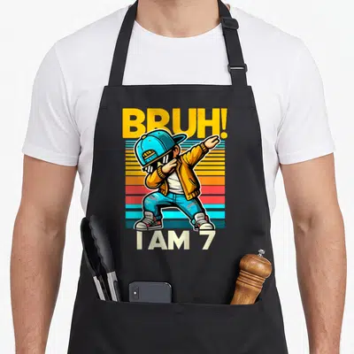 Bruh 7th birthday apron - Cartoon boy dabbing on yellow blue orange red stripes with text BRUH I AM 7 — shown on a black cotton canvas apron against a white background