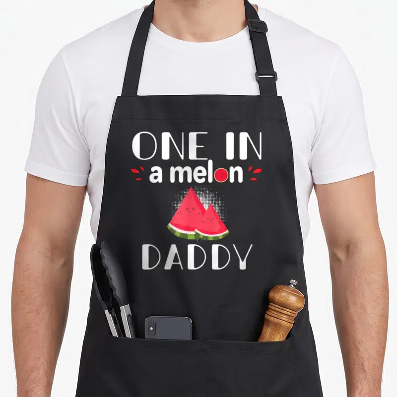 Family watermelon apron - One In a melon DADDY text with two cartoon watermelon slices and red splashes — shown on a black cotton canvas apron against a white background