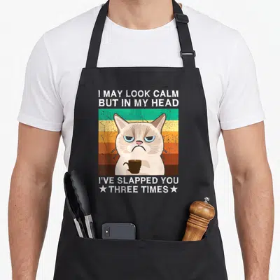Slapped may apron - Grumpy cat holding coffee cup with striped retro background and sarcastic white text — shown on a black cotton canvas apron against a white background