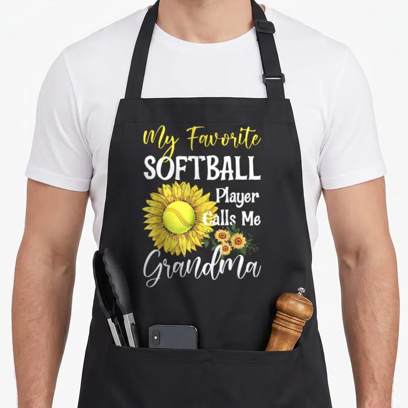 Softball player grandma apron - Stacked text My Favorite Softball Player Calls Me Grandma beside a yellow sunflower — shown on a black cotton canvas apron against a white background