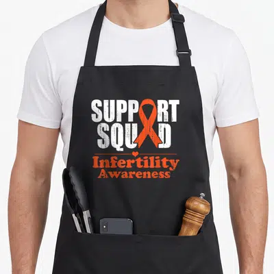 Ivf ribbon apron - SUPPORT SQUAD text with orange awareness ribbon and Infertility Awareness label — shown on a black cotton canvas apron against a white background