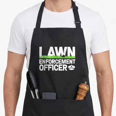 Enforcement landscaper apron - LAWN ENFORCEMENT OFFICER text with grey mower in letters and green grass line — shown on a black cotton canvas apron against a white background