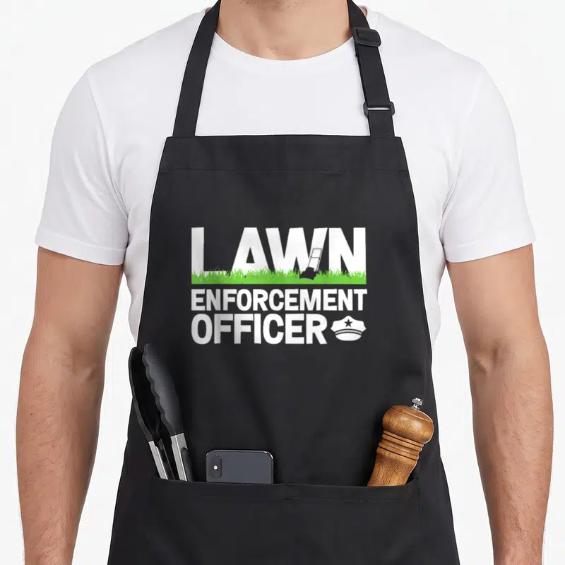 Enforcement landscaper apron - LAWN ENFORCEMENT OFFICER text with grey mower in letters and green grass line — shown on a black cotton canvas apron against a white background