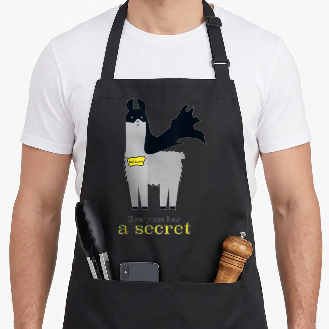 Prob birthday apron - Cartoon grey llama in black mask and cape with yellow BATLLAM tag and secret text — shown on a black cotton canvas apron against a white background