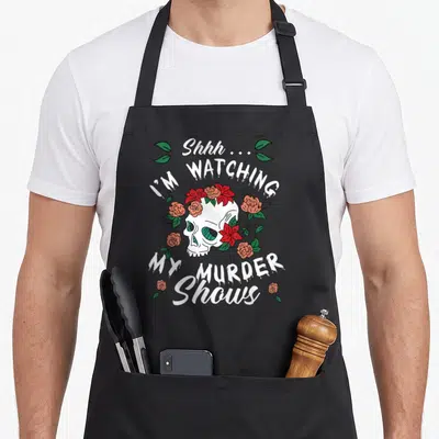 Crime i love watching apron - White skull with red flowers and green leaves surrounded by curved text reading Shhh I'm — shown on a black cotton canvas apron against a white background