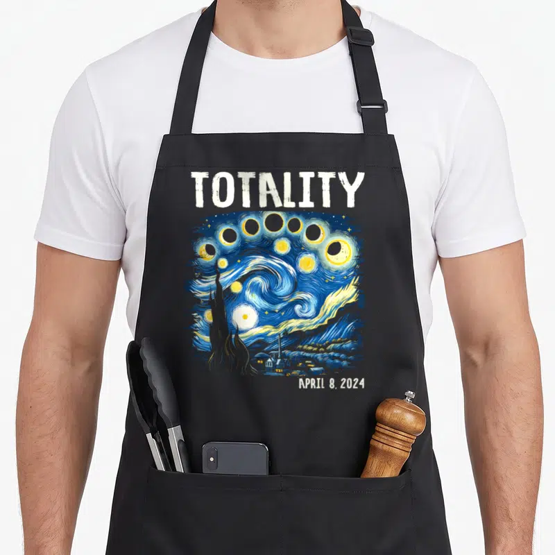 Starry eclipse apron - TOTALITY text above blue swirling sky with moon phases, cypress tree, and village — shown on a black cotton canvas apron against a white background