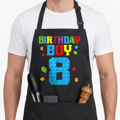 8th grill master apron - Colorful brick letters spelling BIRTHDAY BOY 8 with scattered building blocks — shown on a black cotton canvas apron against a white background