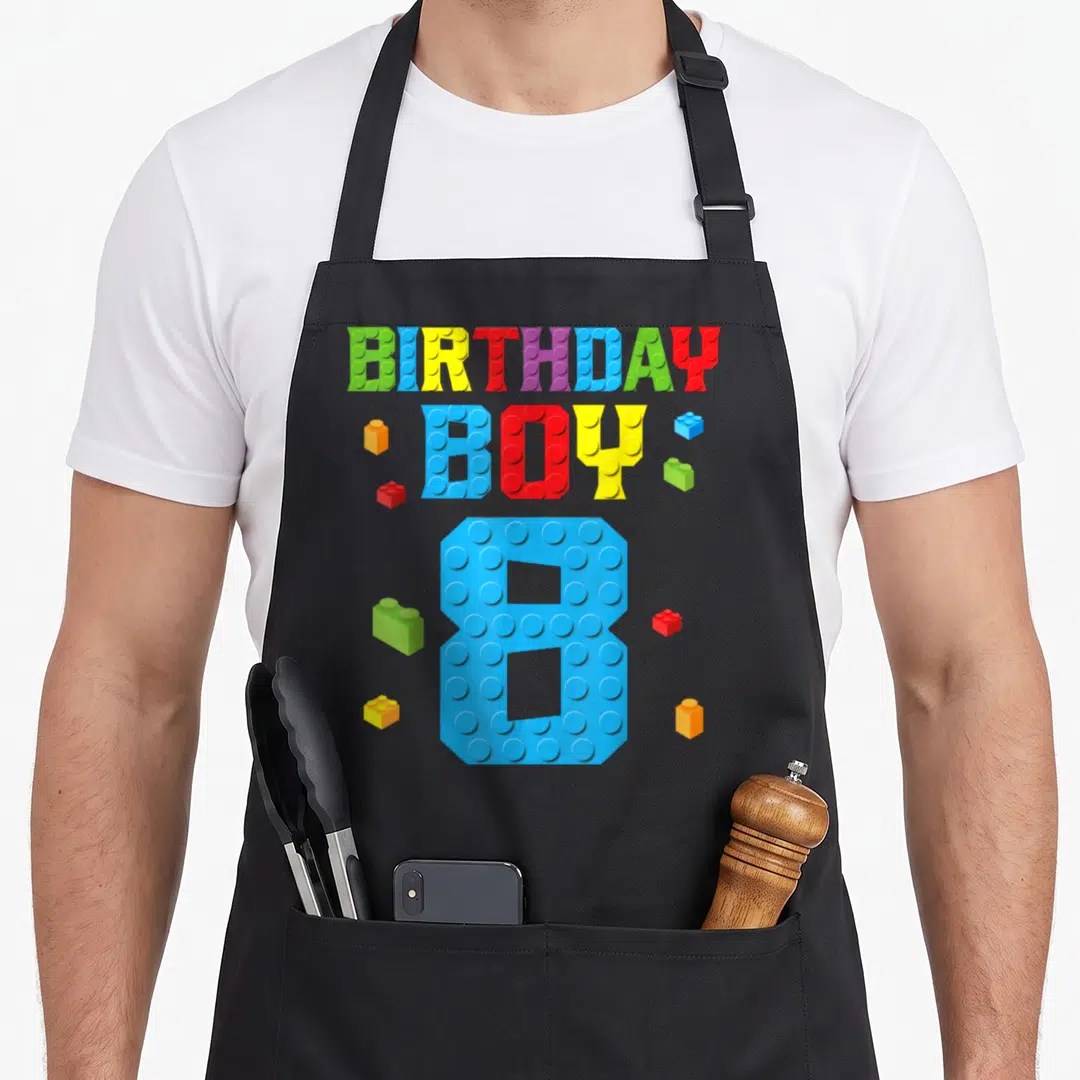 8th grill master apron - Colorful brick letters spelling BIRTHDAY BOY 8 with scattered building blocks — shown on a black cotton canvas apron against a white background