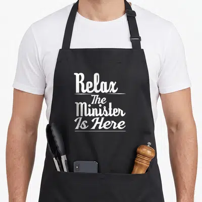 Funny relax minister apron - White stacked text reading Relax The Minister Is Here with horizontal separators — shown on a black cotton canvas apron against a white background