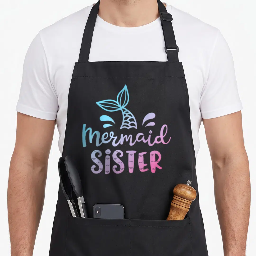 Sister family cooking aprons - Cartoon mermaid tail and splashes above stacked text reading mermaid SISTER in blue — shown on a black cotton canvas apron against a white background