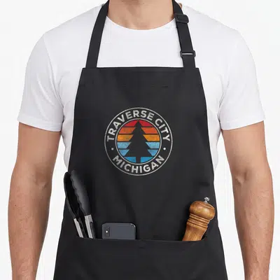 Traverse 70s apron - Retro circular badge with pine tree silhouette over red orange and blue stripes and white text — shown on a black cotton canvas apron against a white background