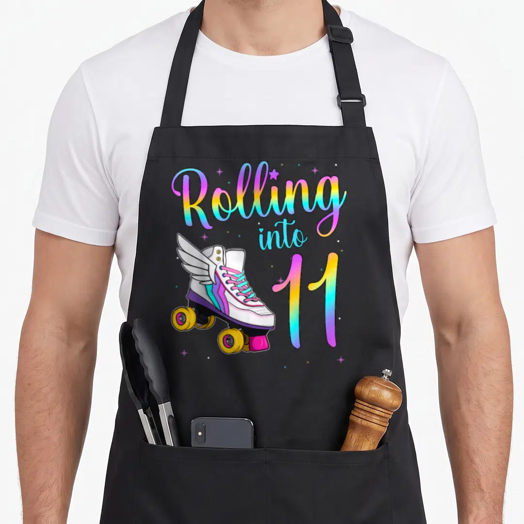 Rolling roll roller skate turning apron - Winged roller skate with rainbow 'Rolling into 11' text and scattered stars — shown on a black cotton canvas apron against a white background