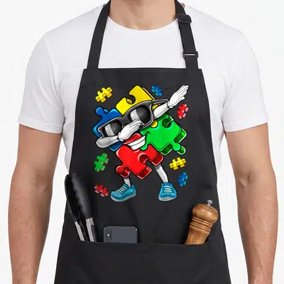 Autism puzzles apron - Cartoon dabbing character made of yellow, blue, red, green, and white puzzle pieces wearing — shown on a black cotton canvas apron against a white background