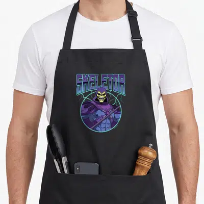 Skeletor lighting bolt apron - Arched SKELETOR text above character in purple blue circle with lightning bolts — shown on a black cotton canvas apron against a white background
