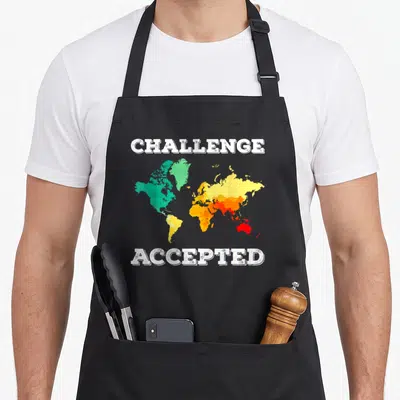 Challenge travel aprons - Distressed world map in green yellow orange red with Challenge Accepted text above and below — shown on a black cotton canvas apron against a white background