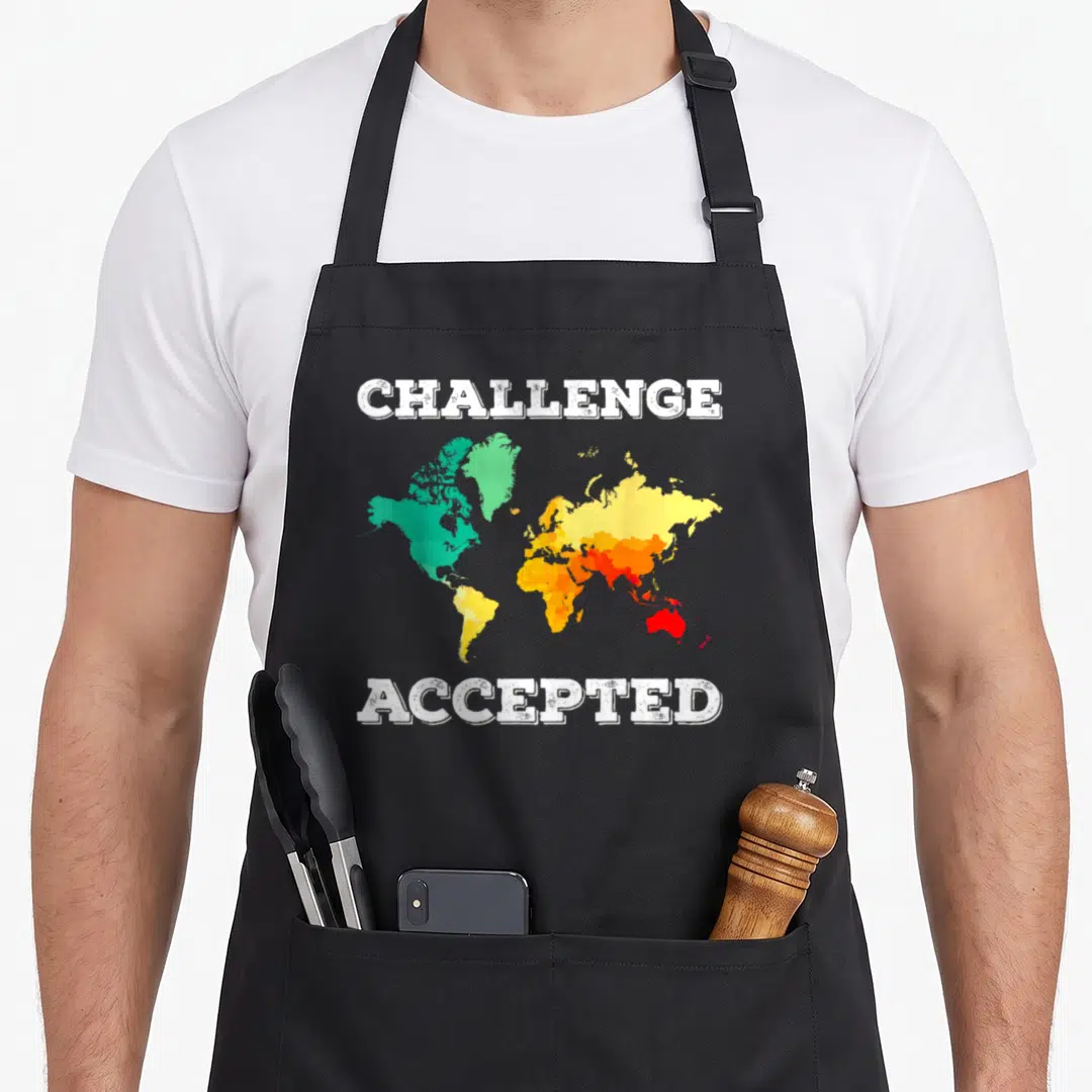 Challenge travel aprons - Distressed world map in green yellow orange red with Challenge Accepted text above and below — shown on a black cotton canvas apron against a white background