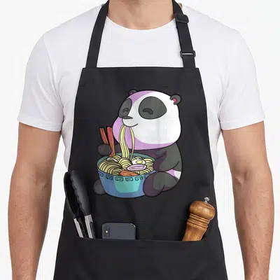 Panda kawaii cooking apron - Cartoon panda sitting and eating yellow noodles from a blue bowl with chopsticks — shown on a black cotton canvas apron against a white background