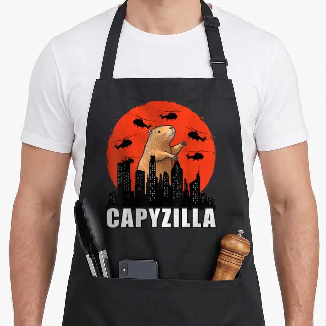 Rodent cute capybara apron - Giant brown capybara behind black skyline with red sun and five helicopters — shown on a black cotton canvas apron against a white background