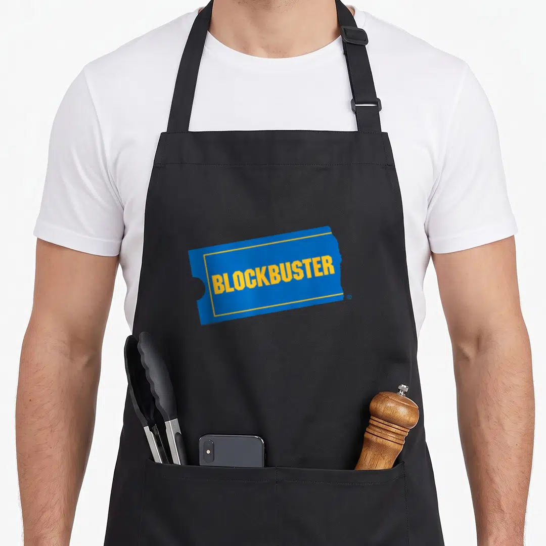Blockbuster video lover apron - Blue ticket shape with yellow BLOCKBUSTER text centered on black fabric — shown on a black cotton canvas apron against a white background