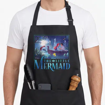 Eric mermaid apron - Ariel Eric boat scene with Sebastian Flounder and The Little Mermaid title text in blue — shown on a black cotton canvas apron against a white background