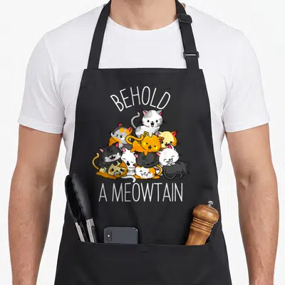 Behold kitty cat apron - Cartoon cats in white, orange, grey, black piled under arched BEHOLD A MEOWTAIN text — shown on a black cotton canvas apron against a white background