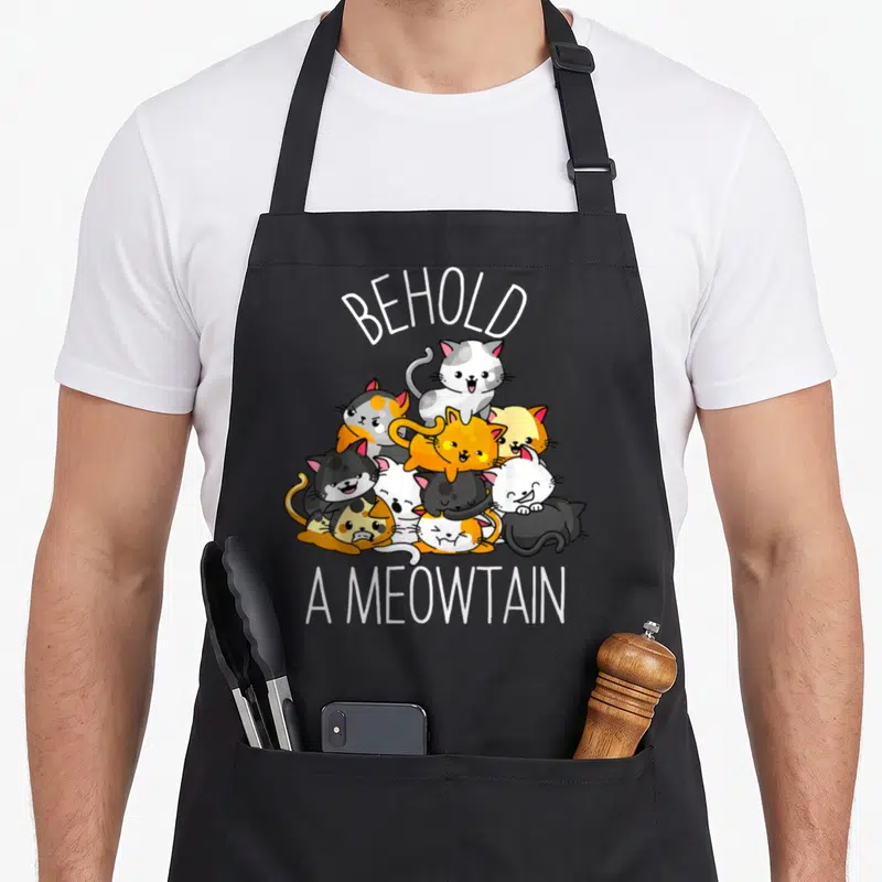 Behold kitty cat apron - Cartoon cats in white, orange, grey, black piled under arched BEHOLD A MEOWTAIN text — shown on a black cotton canvas apron against a white background