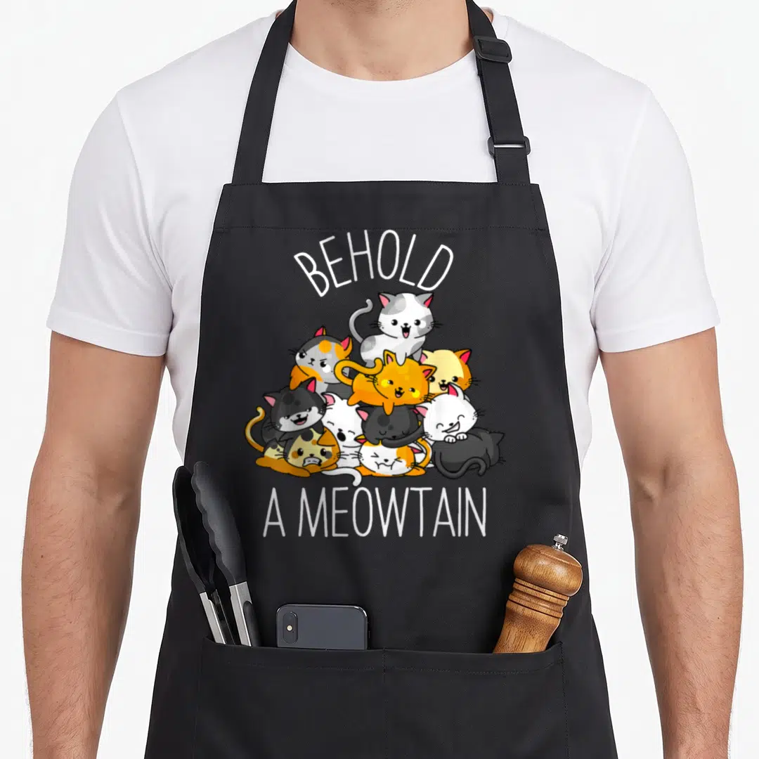 Behold kitty cat apron - Cartoon cats in white, orange, grey, black piled under arched BEHOLD A MEOWTAIN text — shown on a black cotton canvas apron against a white background