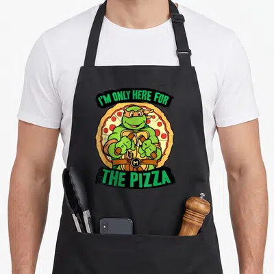Funny pizza apron - Green turtle holding nunchunks in front of pepperoni pizza with arched text banners — shown on a black cotton canvas apron against a white background