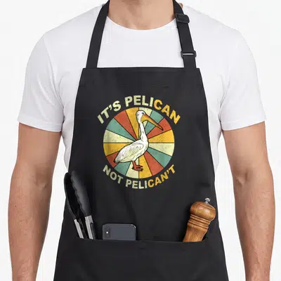 Pelicant funny pelican lover apron - White pelican in front of yellow red green sunburst with arched retro text — shown on a black cotton canvas apron against a white background