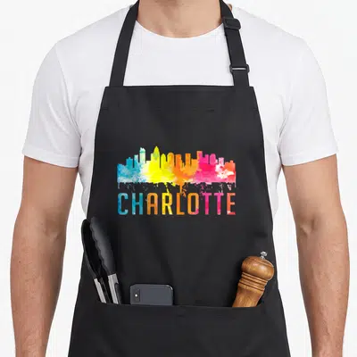 Carolina charlotte apron - Watercolor city skyline in blue, yellow, pink, and orange above the word CHARLOTTE — shown on a black cotton canvas apron against a white background