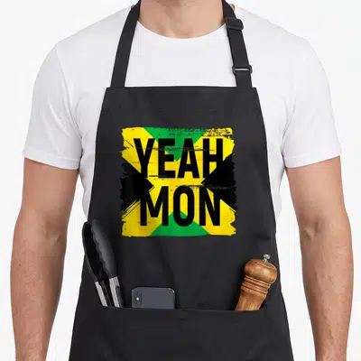 Yeah pride apron - Distressed yellow green and black Jamaican flag with YEAH MON text overlay — shown on a black cotton canvas apron against a white background