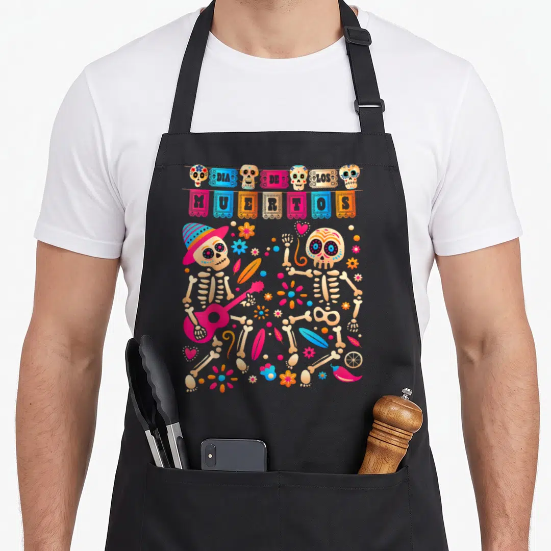 Mexican day of the dead apron - DIA DE LOS MUERTOS banner above two skeletons and colorful flowers — shown on a black cotton canvas apron against a white background