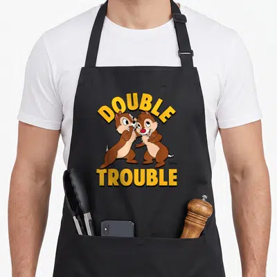 Chip double apron - Two brown chipmunks with yellow arched DOUBLE TROUBLE text in cartoon style — shown on a black cotton canvas apron against a white background