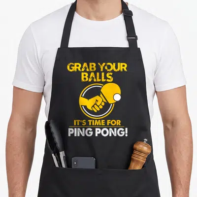Grab table tennis apron - Yellow and white cartoon paddle graphic with GRAB YOUR BALLS IT'S TIME FOR PING PONG text — shown on a black cotton canvas apron against a white background
