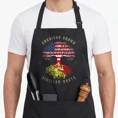 Root sicily apron - Tree with US flag canopy and Triskelion root symbol bearing American Grown Sicilian Roots text — shown on a black cotton canvas apron against a white background