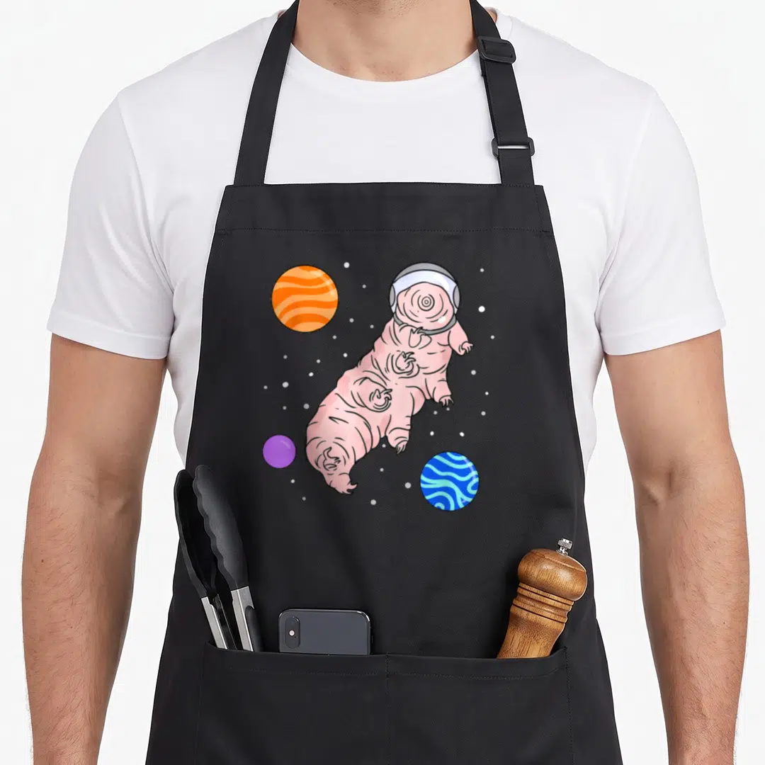 Space microbiologist apron - Pink tardigrade in space helmet floating near orange, blue, and purple planets with white — shown on a black cotton canvas apron against a white background