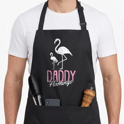 Flamingo father day apron - Two white flamingos above pink DADDY and white Flamingo text on black fabric — shown on a black cotton canvas apron against a white background