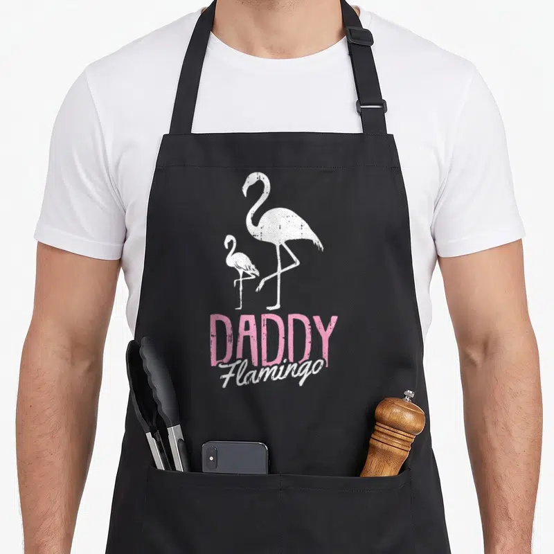 Flamingo father day apron - Two white flamingos above pink DADDY and white Flamingo text on black fabric — shown on a black cotton canvas apron against a white background