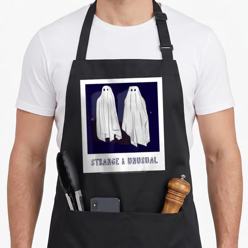 Polaroid ghost apron - Two white cartoon ghosts in dark blue square above STRANGE & UNUSUAL text — shown on a black cotton canvas apron against a white background