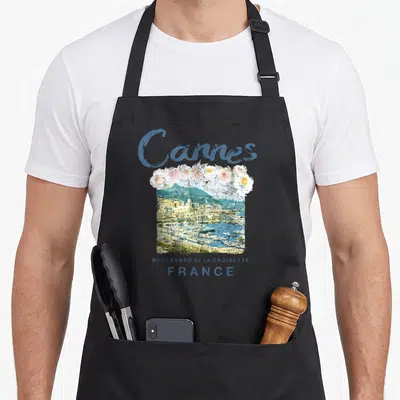 Canne french apron - Cannes script, pink flowers, harbor photo, Boulevard de la Croisette text — shown on a black cotton canvas apron against a white background